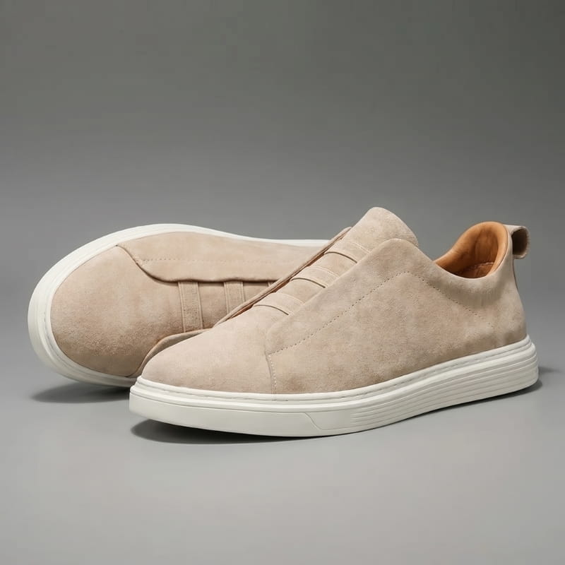 Pair of beige suede slip-on sneakers with white soles facing forward on a gray studio background