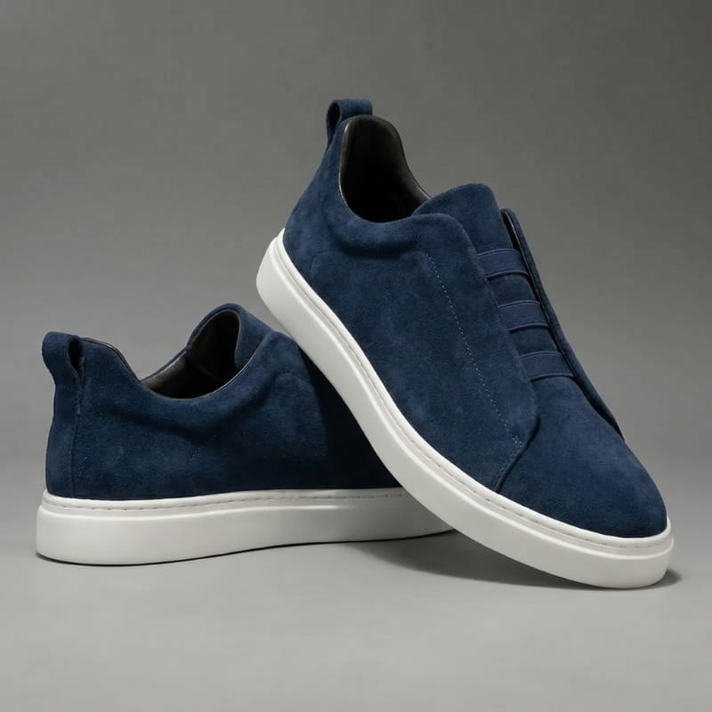 Pair of navy blue suede slip-on sneakers with white soles stacked at an angle on a gray studio background