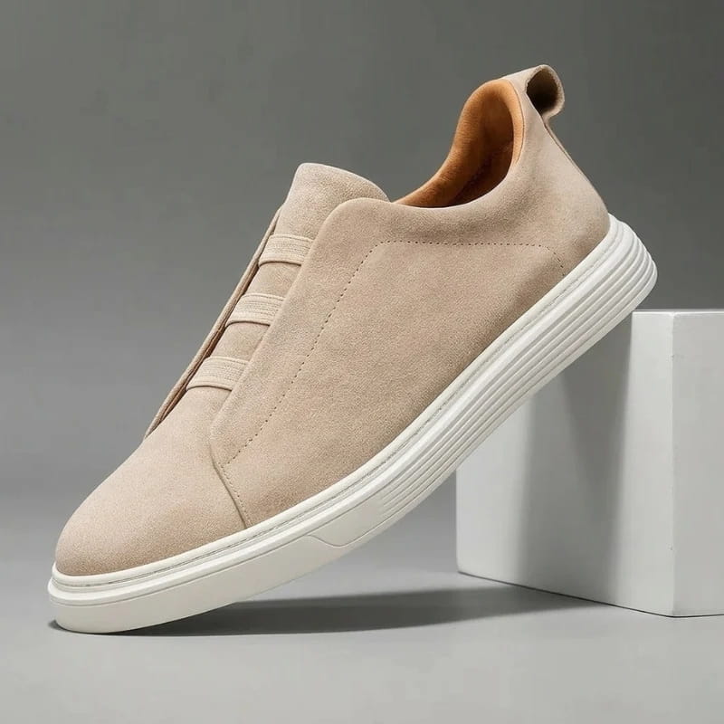 Beige suede slip-on sneaker with elastic stretch-lace panel and white sole propped diagonally against a white block