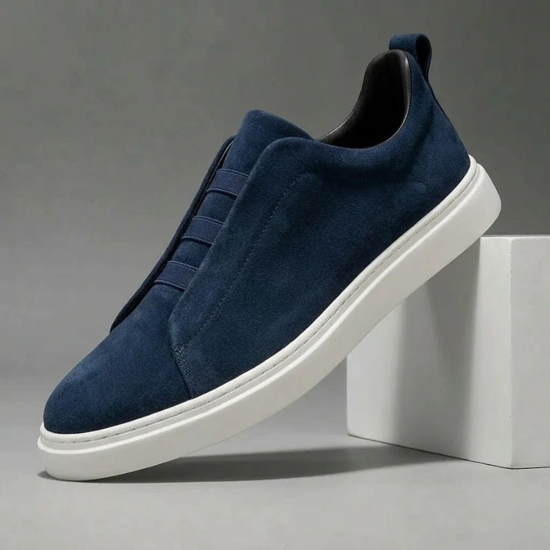 Navy blue suede slip-on sneaker with elastic stretch-lace panel and white sole propped diagonally against a white block
