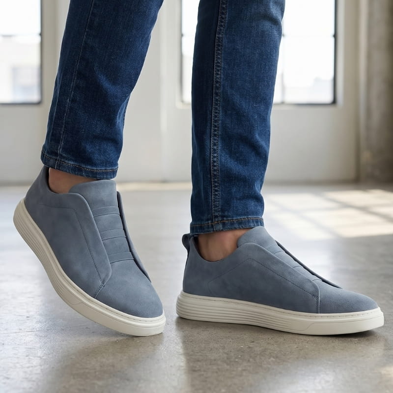 Person wearing blue-gray suede slip-on sneakers with jeans, walking indoors in a bright sunlit space