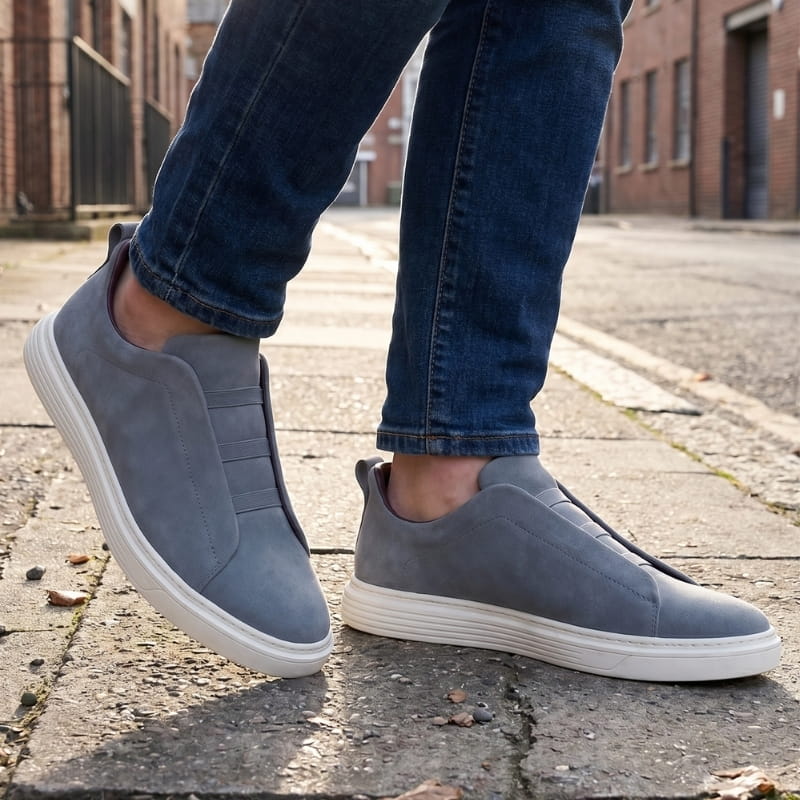 Person wearing blue-gray suede slip-on sneakers with jeans, walking on a cobblestone street with fallen leaves