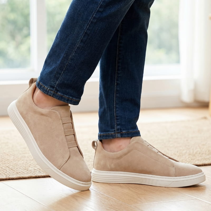 Person wearing beige suede slip-on sneakers with jeans, walking on a wood floor near a rug indoors
