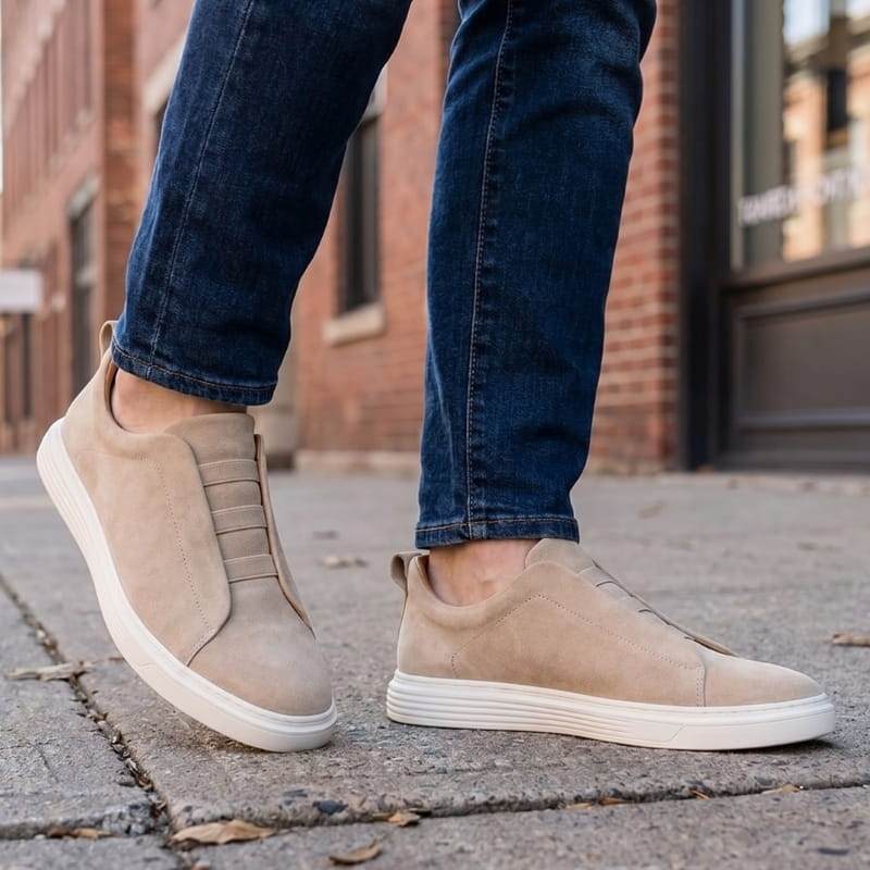 Person wearing beige suede slip-on sneakers with jeans, walking on a paved street lined with brick buildings