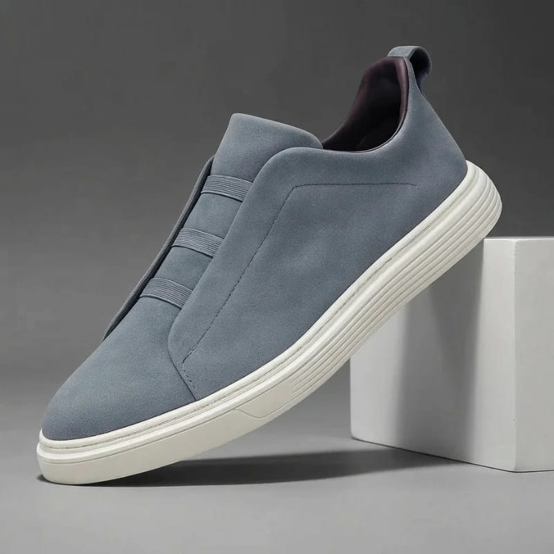 Blue-gray suede slip-on sneaker with elastic stretch-lace panel and white sole propped diagonally against a white block