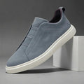 Blue-gray suede slip-on sneaker with elastic stretch-lace panel and white sole propped diagonally against a white block