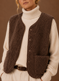 Three-quarter view of a woman wearing a brown fleece sweater vest with front patch pockets over a cream turtleneck sweater.