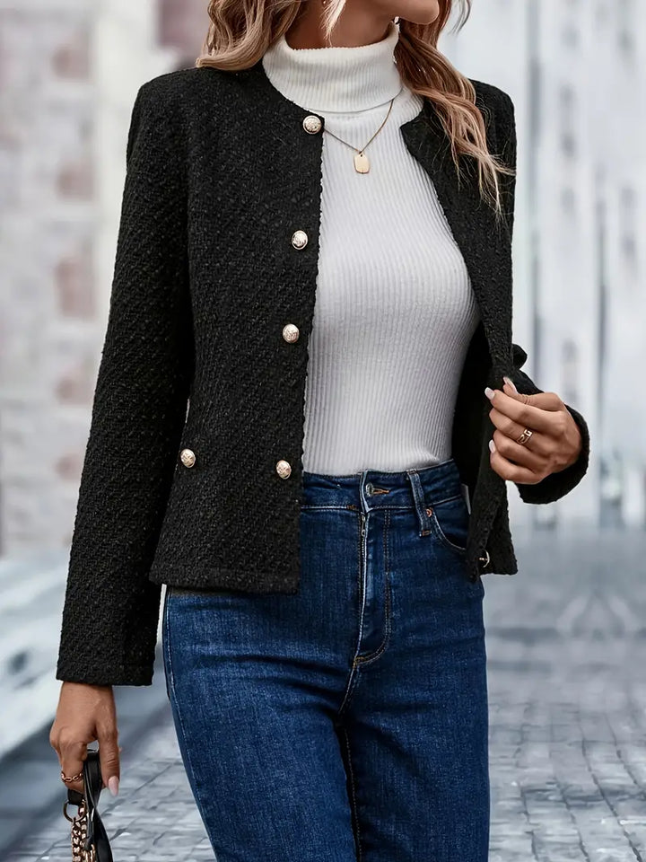 Woman wearing a black textured tweed jacket with gold-tone buttons over a white ribbed sweater and blue jeans while walking outdoors.