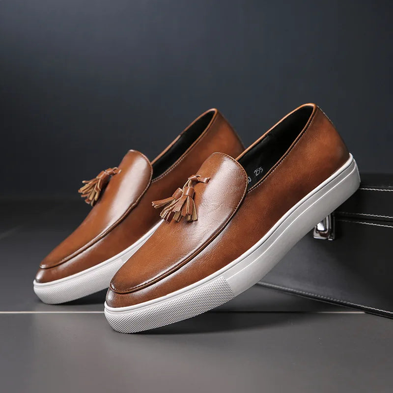 Pair of brown leather tassel loafers with white soles, one propped against a dark leather bag, against a gray backdrop.