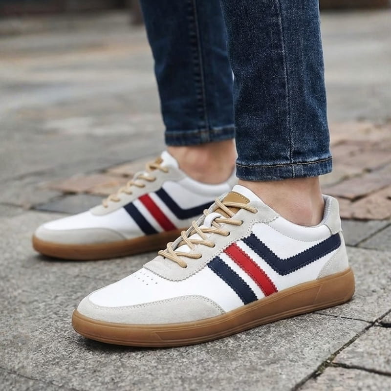 Person wearing white leather retro court sneakers with navy and red stripes and jeans, standing on a wet cobblestone street