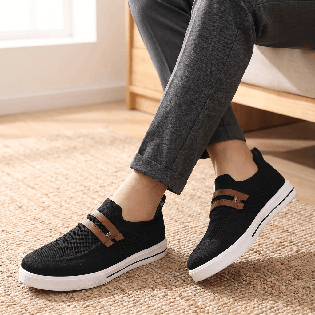 Close-up of crossed ankles wearing gray trousers and black mesh Orion sneakers with brown straps, on a jute rug near a window