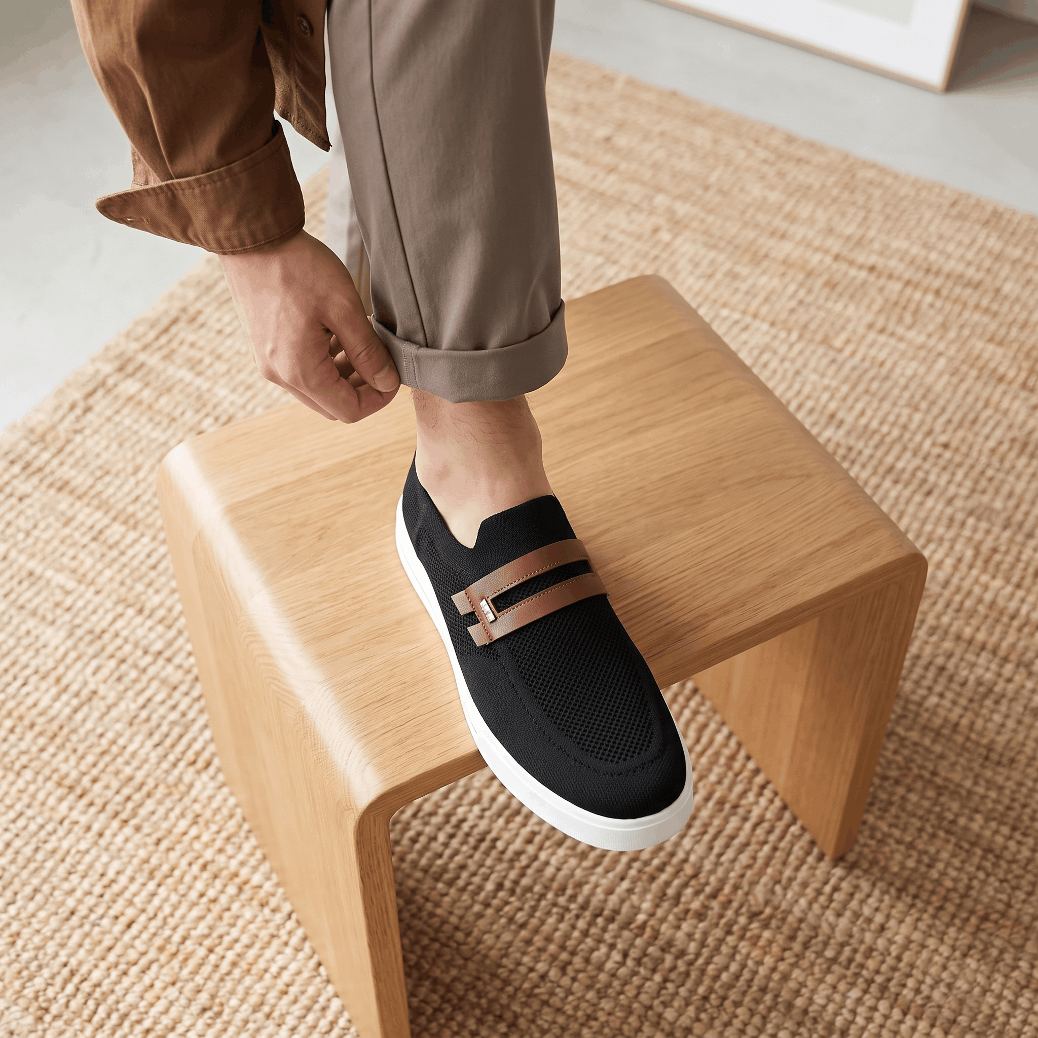 Man adjusting trouser cuff with foot on a wooden stool wearing black mesh Orion sneaker, on a jute rug