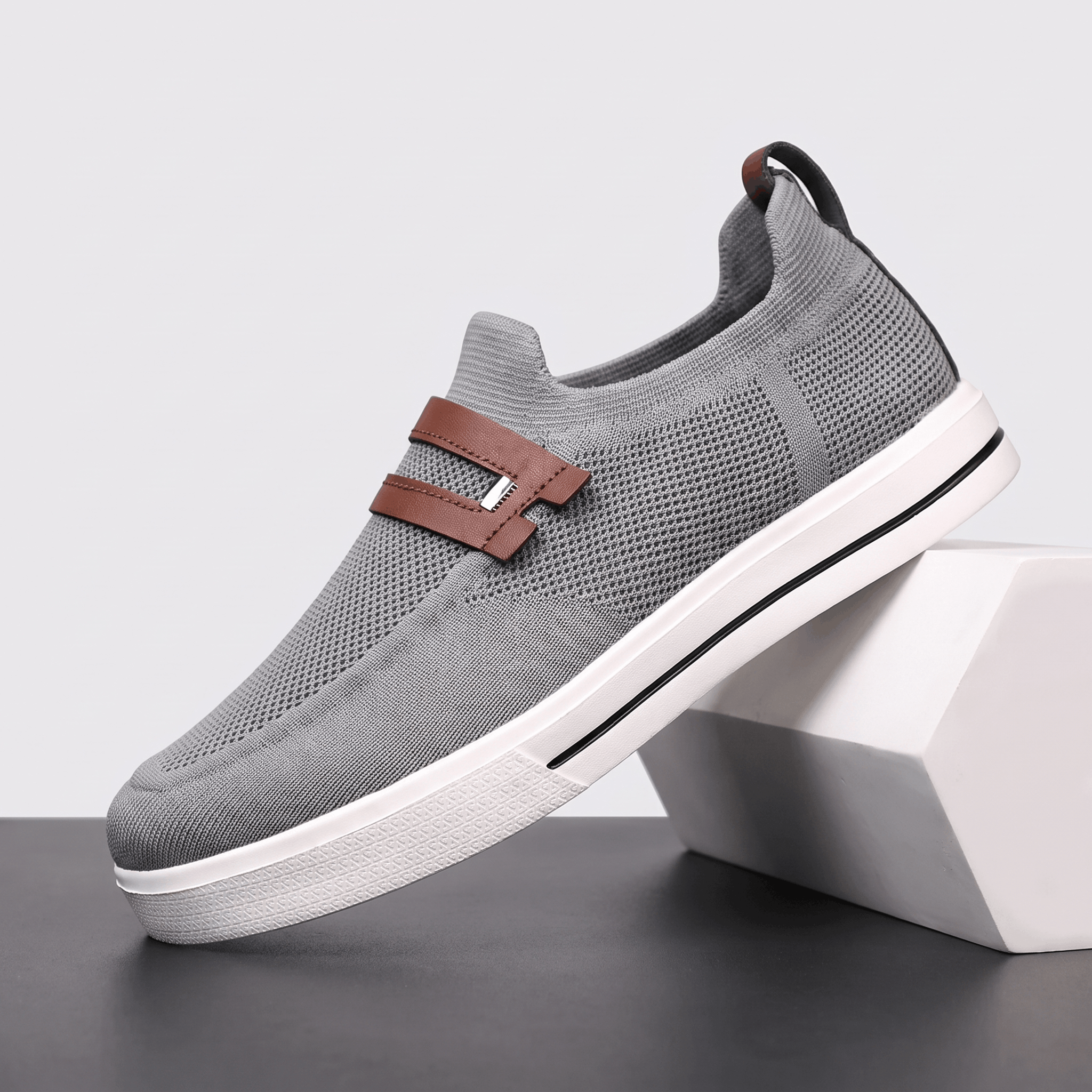 Single gray mesh Orion slip-on sneaker with brown leather strap, propped against a white block against a studio backdrop