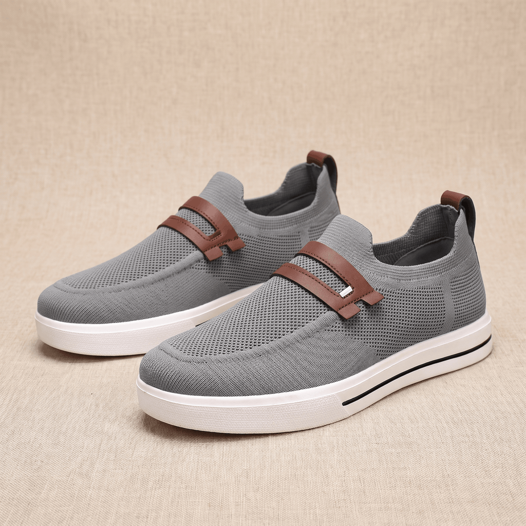 Pair of gray mesh Orion slip-on sneakers with brown leather straps, resting on a beige fabric backdrop