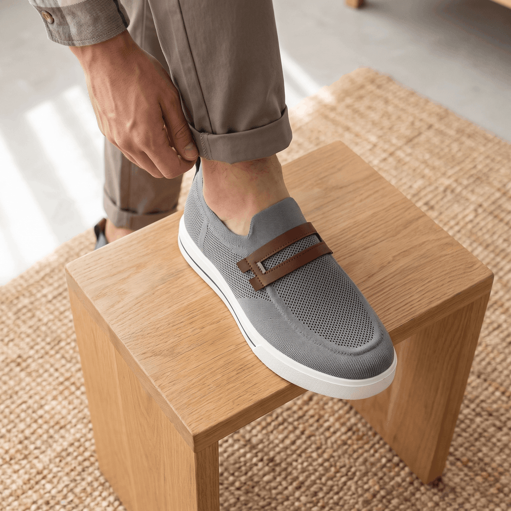Man adjusting trouser cuff with foot resting on a wooden block wearing gray mesh Orion sneaker, on a jute rug