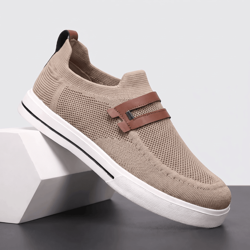 Single khaki mesh Orion slip-on sneaker with brown leather strap, propped against a white block against a studio backdrop