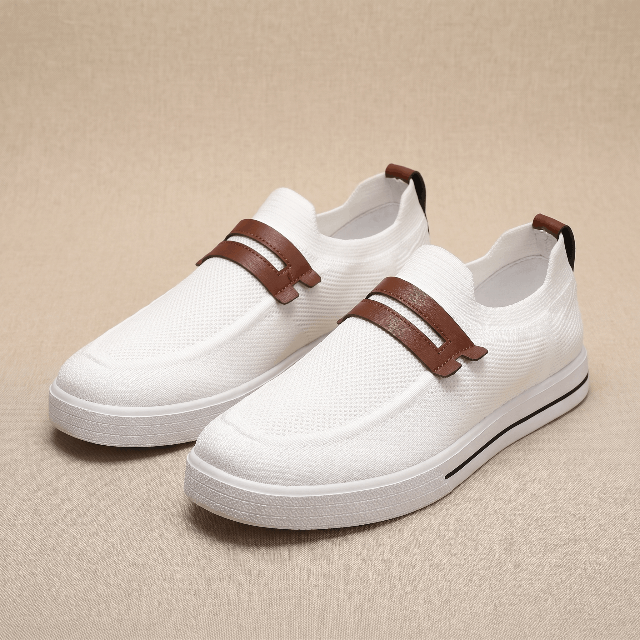 Pair of white mesh Orion slip-on sneakers with brown leather straps, resting on a beige fabric backdrop