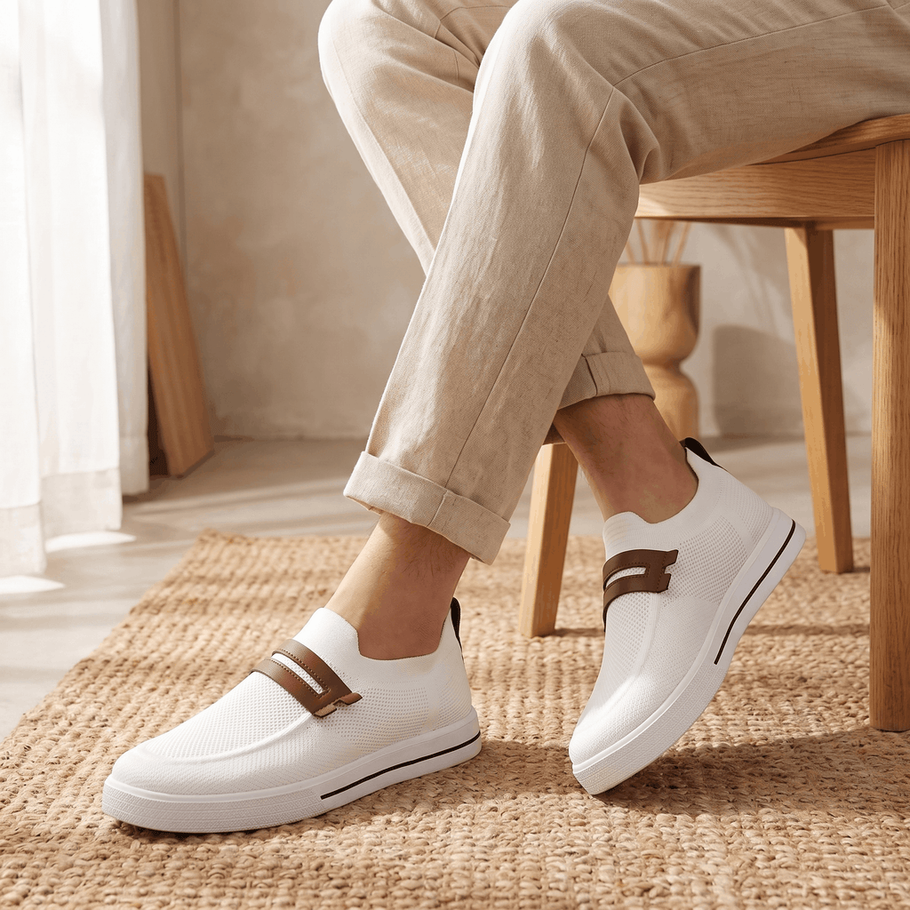 Close-up of legs wearing beige trousers and white mesh Orion sneakers with brown straps, on a jute rug near a wooden chair