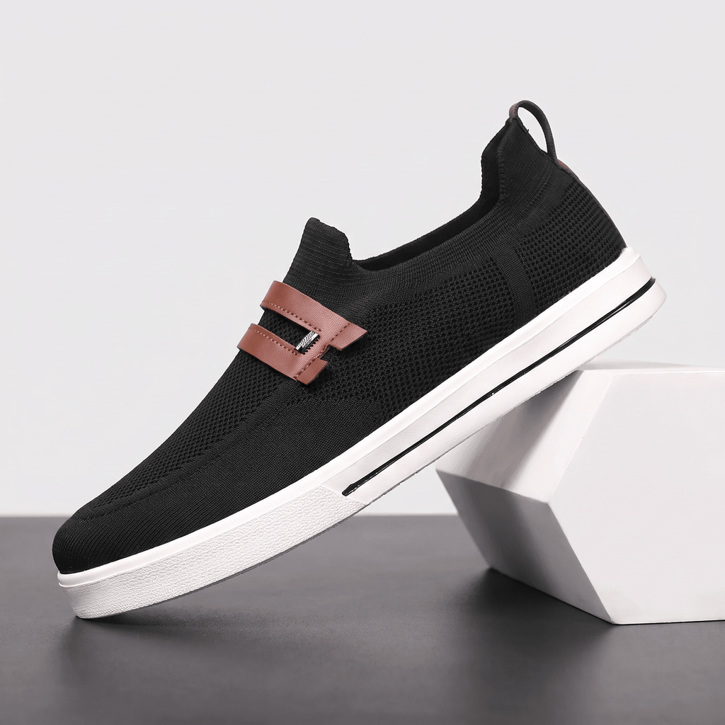 Single black mesh Orion slip-on sneaker with brown leather strap, propped against a white block against a studio backdrop