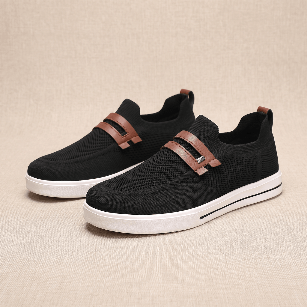 Pair of black mesh Orion slip-on sneakers with brown leather straps, resting on a beige fabric backdrop