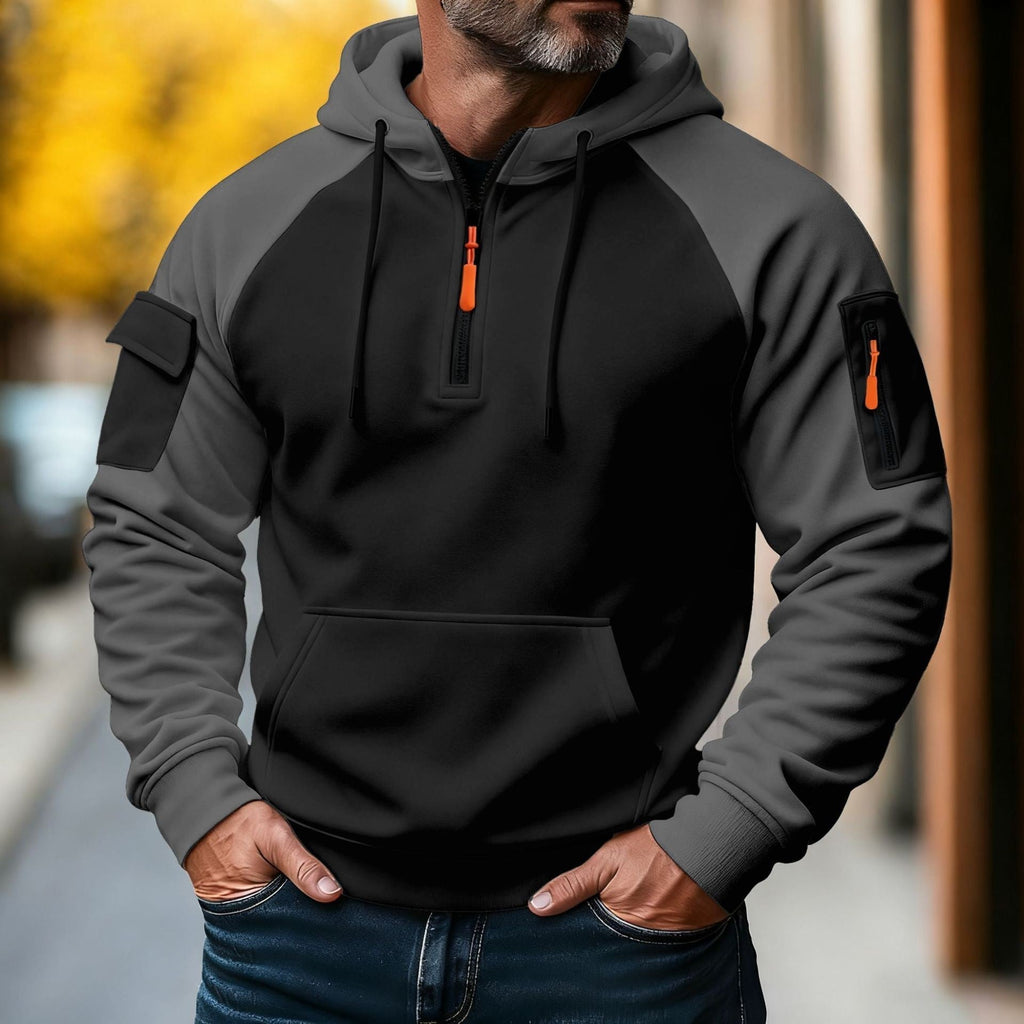 Man wearing a black and charcoal color-block tactical hoodie with a half-zip neckline, adjustable hood, utility sleeve pocket, and kangaroo pocket.