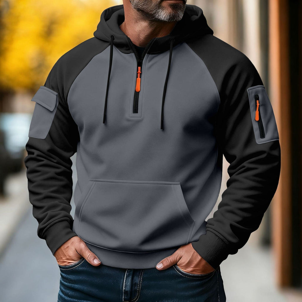 Man wearing a charcoal and black color-block tactical hoodie featuring a half-zip front, adjustable hood, sleeve zip pocket, and kangaroo pocket.