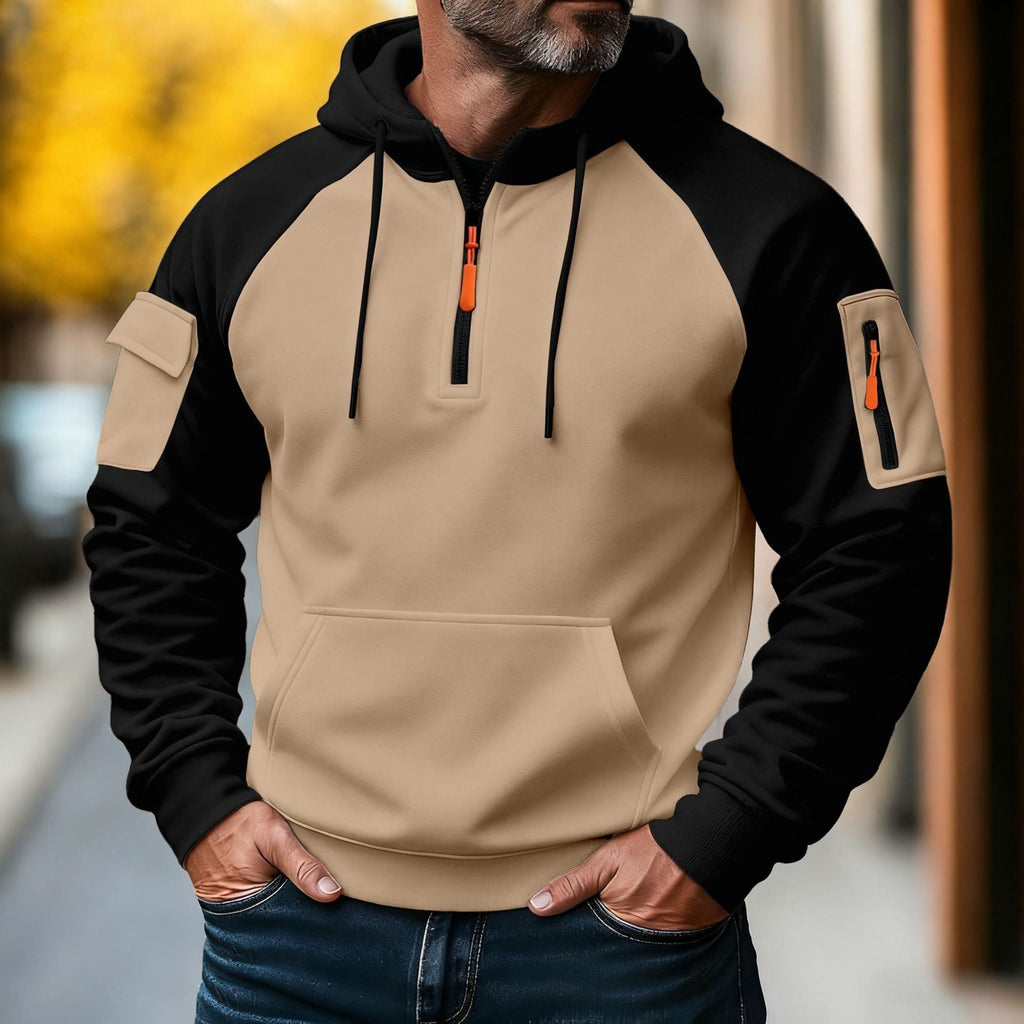 Man wearing a khaki and black color-block tactical hoodie with an adjustable hood, half-zip neckline, utility sleeve pocket, and front kangaroo pocket.