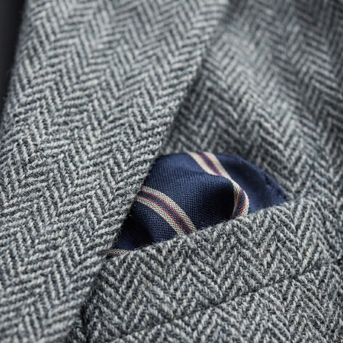 Close-up of the chest pocket featuring a patterned pocket square on a men's grey herringbone tweed blazer.
