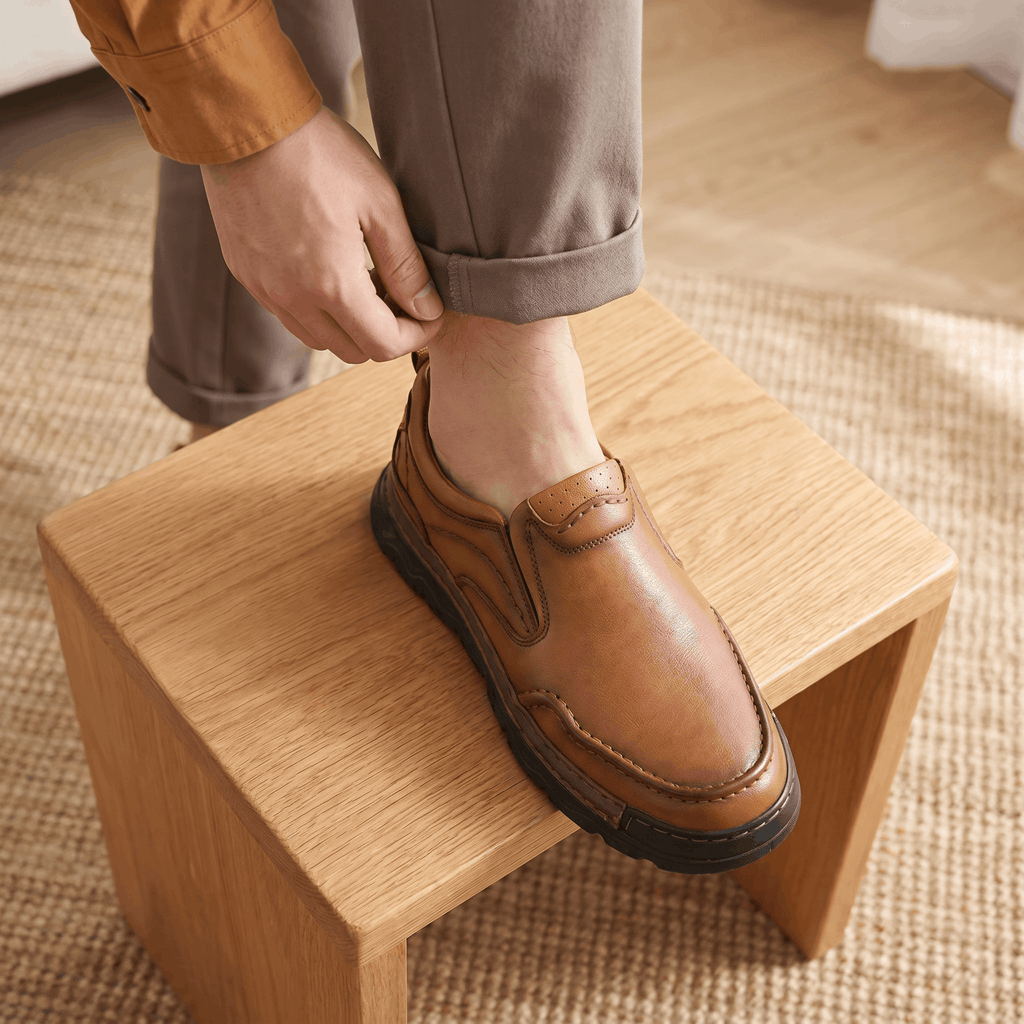 Man adjusting trouser cuff with foot resting on a wooden block wearing brown leather Piolo loafer, on a jute rug