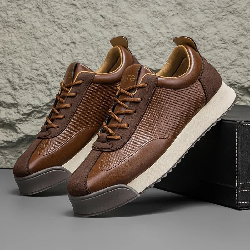 Pair of brown leather sneakers with cream soles, one propped against a black shoe box, against a textured stone backdrop.