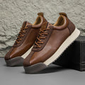 Pair of brown leather sneakers with cream soles, one propped against a black shoe box, against a textured stone backdrop.