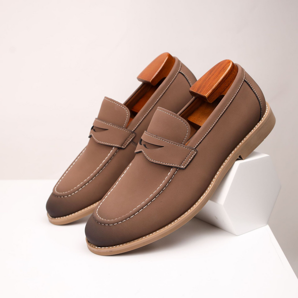 Pair of taupe leather penny loafers with wooden shoe trees, resting against a small white block on a white surface.