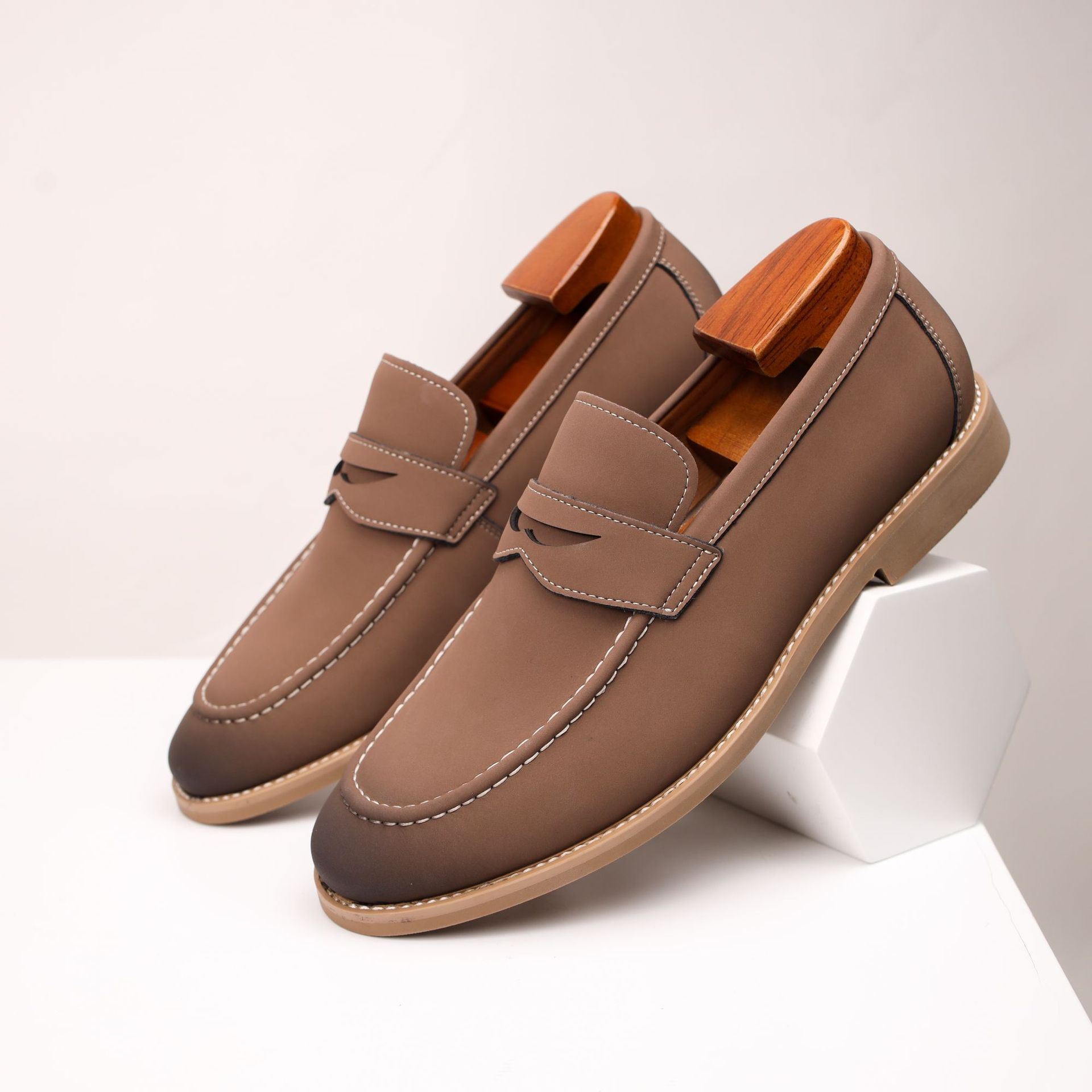 Pair of taupe leather penny loafers with wooden shoe trees, resting against a small white block on a white surface.