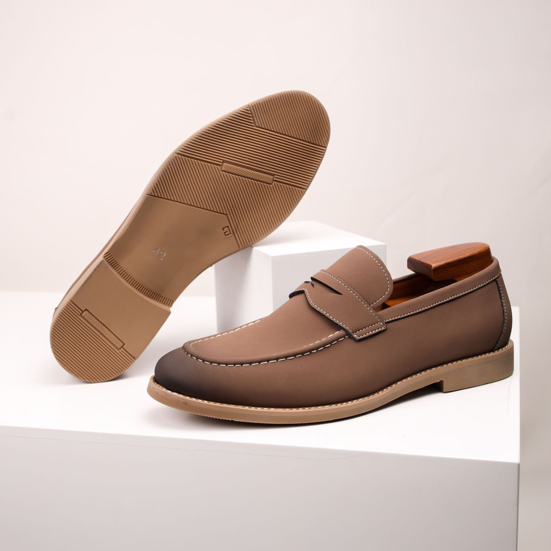 A brown leather penny loafer with a wooden shoe tree standing upright, beside another tilted on its side to show the tread sole, on white blocks.