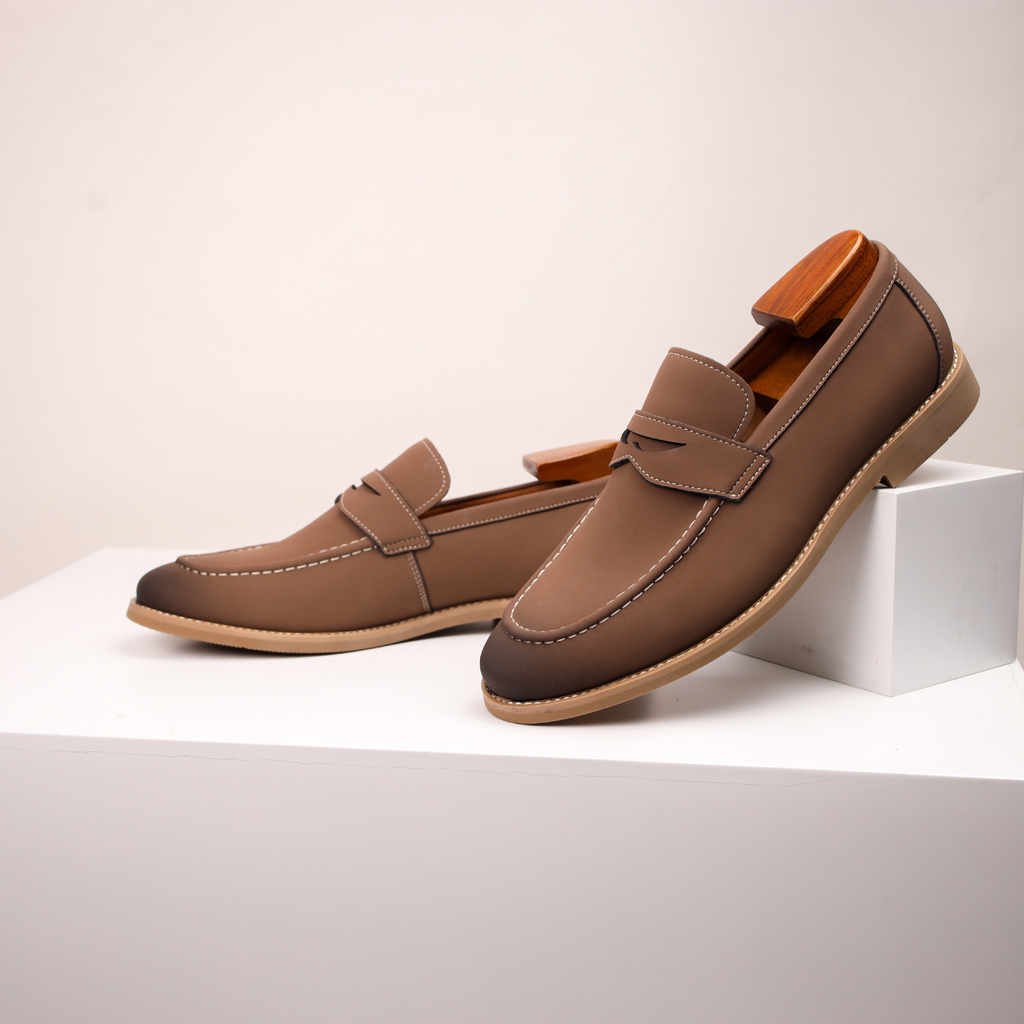 Pair of brown leather penny loafers with a wooden shoe tree, one propped against a small white block on a white surface.