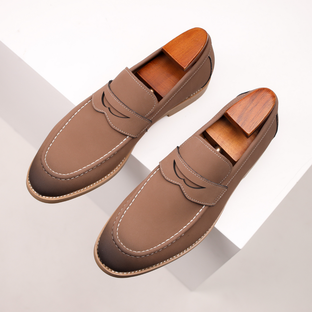 Overhead view of a pair of taupe leather penny loafers with wooden shoe trees, resting on angled white geometric blocks.