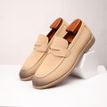 Pair of sand-colored suede penny loafers with wooden shoe trees, one propped against a small white block on a white surface.