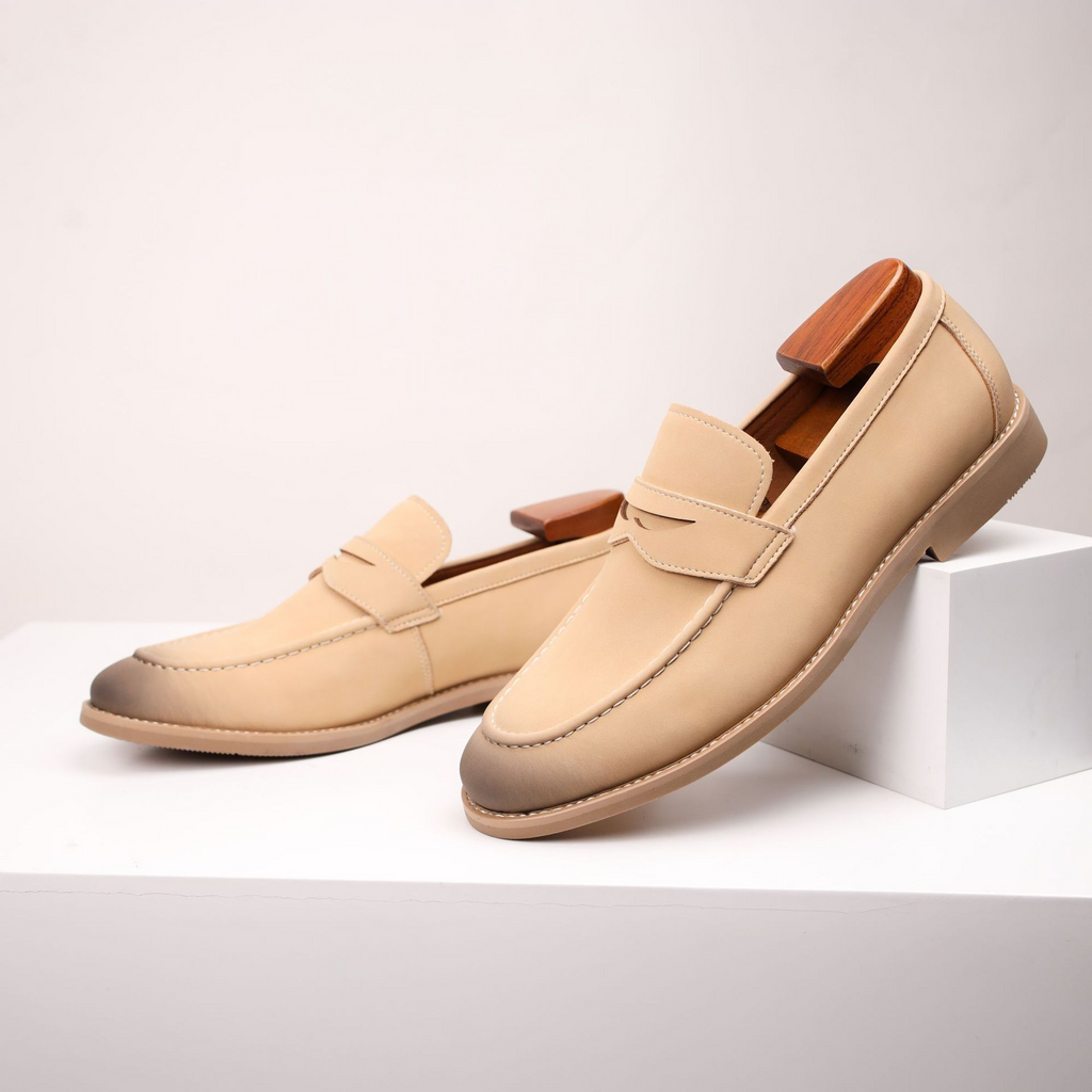 Close-up side view of sand-colored suede penny loafers with wooden shoe trees, resting against a small white block.