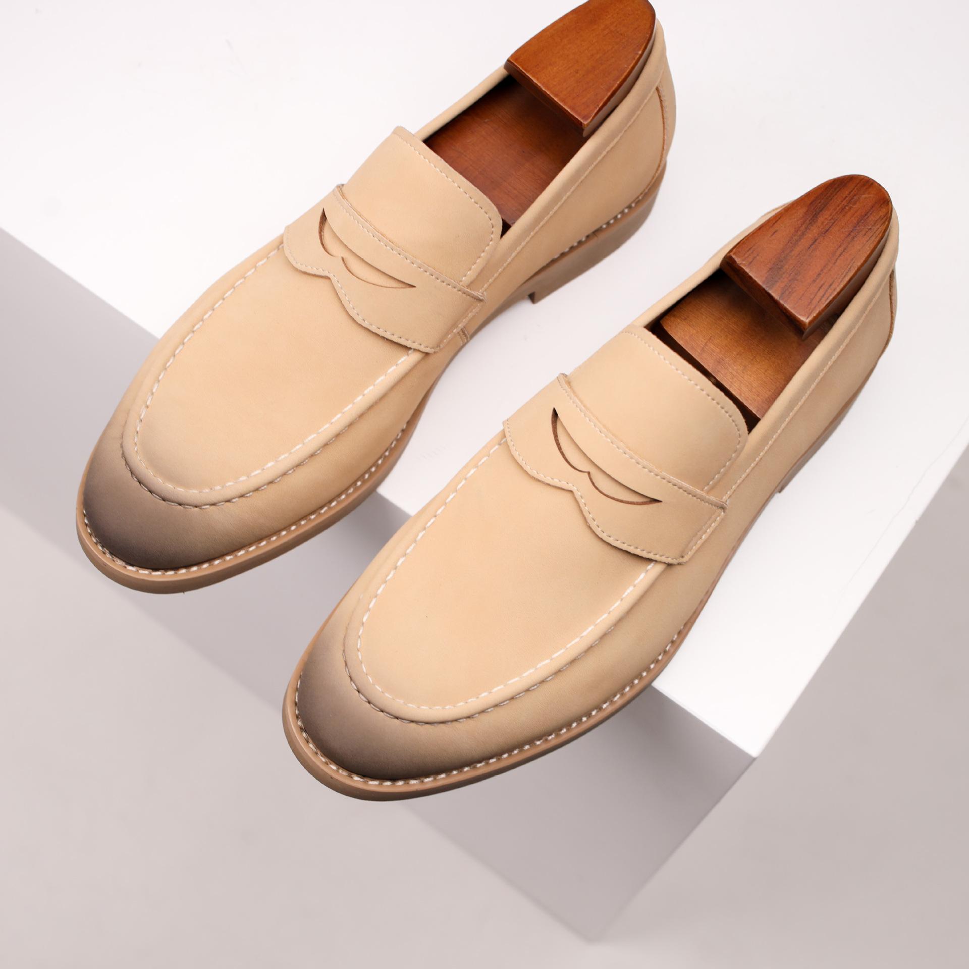 Overhead angled view of a pair of tan leather penny loafers with burnished toes, resting on a white geometric platform.