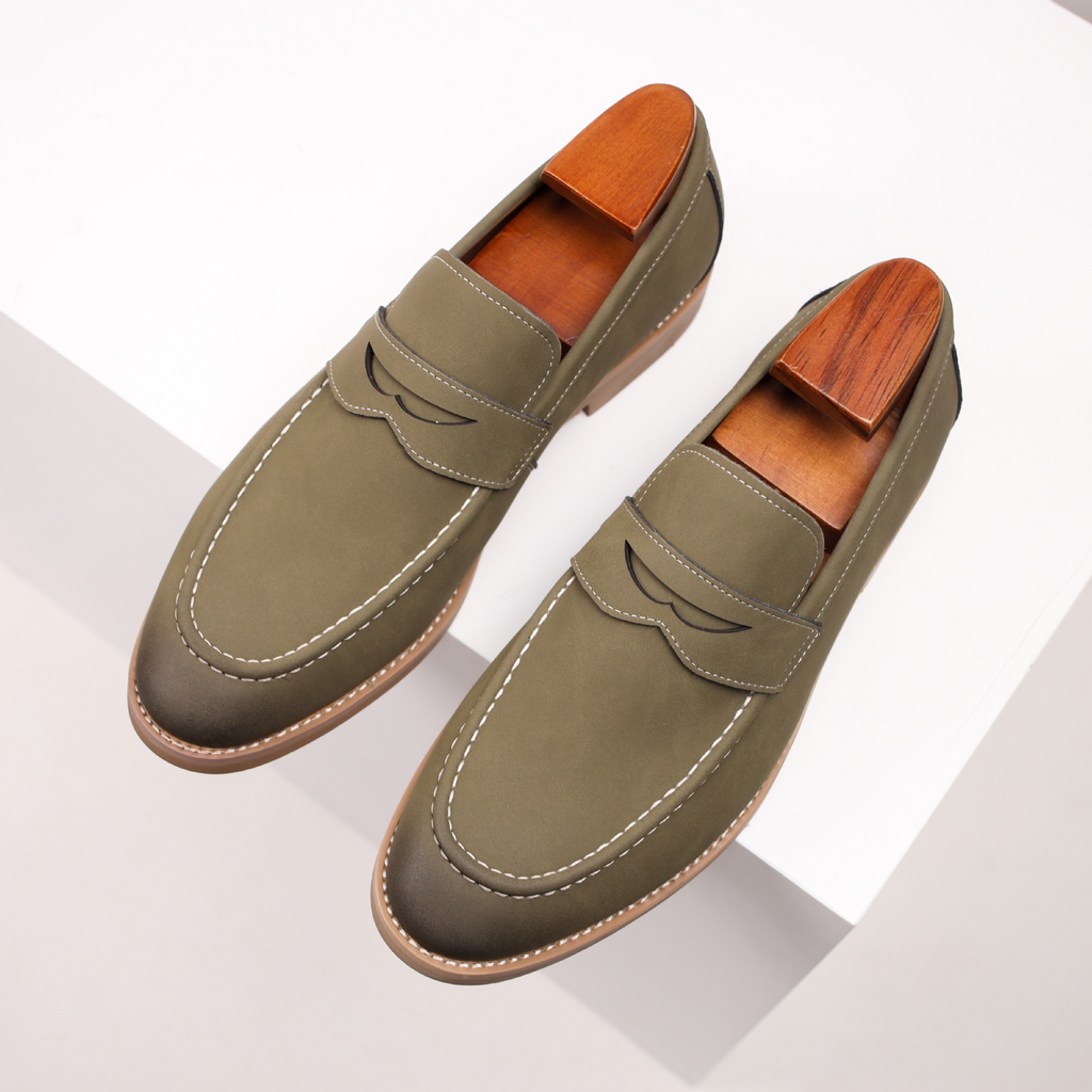 Overhead angled view of a pair of olive green leather penny loafers, resting on a white geometric platform.