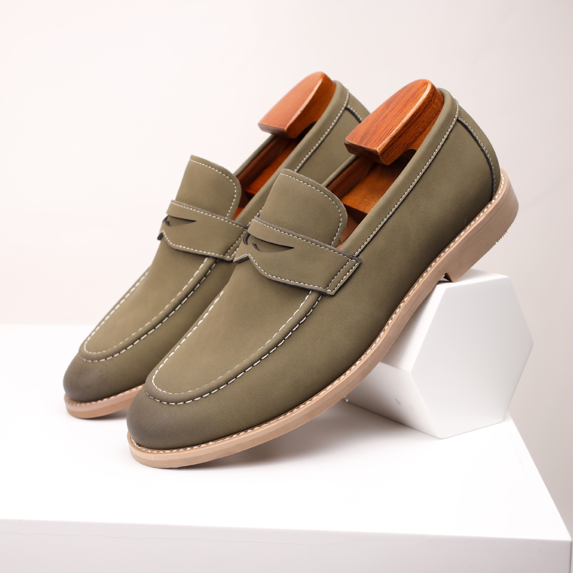 Pair of olive green leather penny loafers standing beside a white geometric block on a white pedestal.