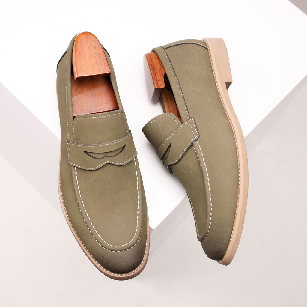 Overhead view of a pair of olive green leather penny loafers with wooden shoe trees, angled on a white geometric platform.