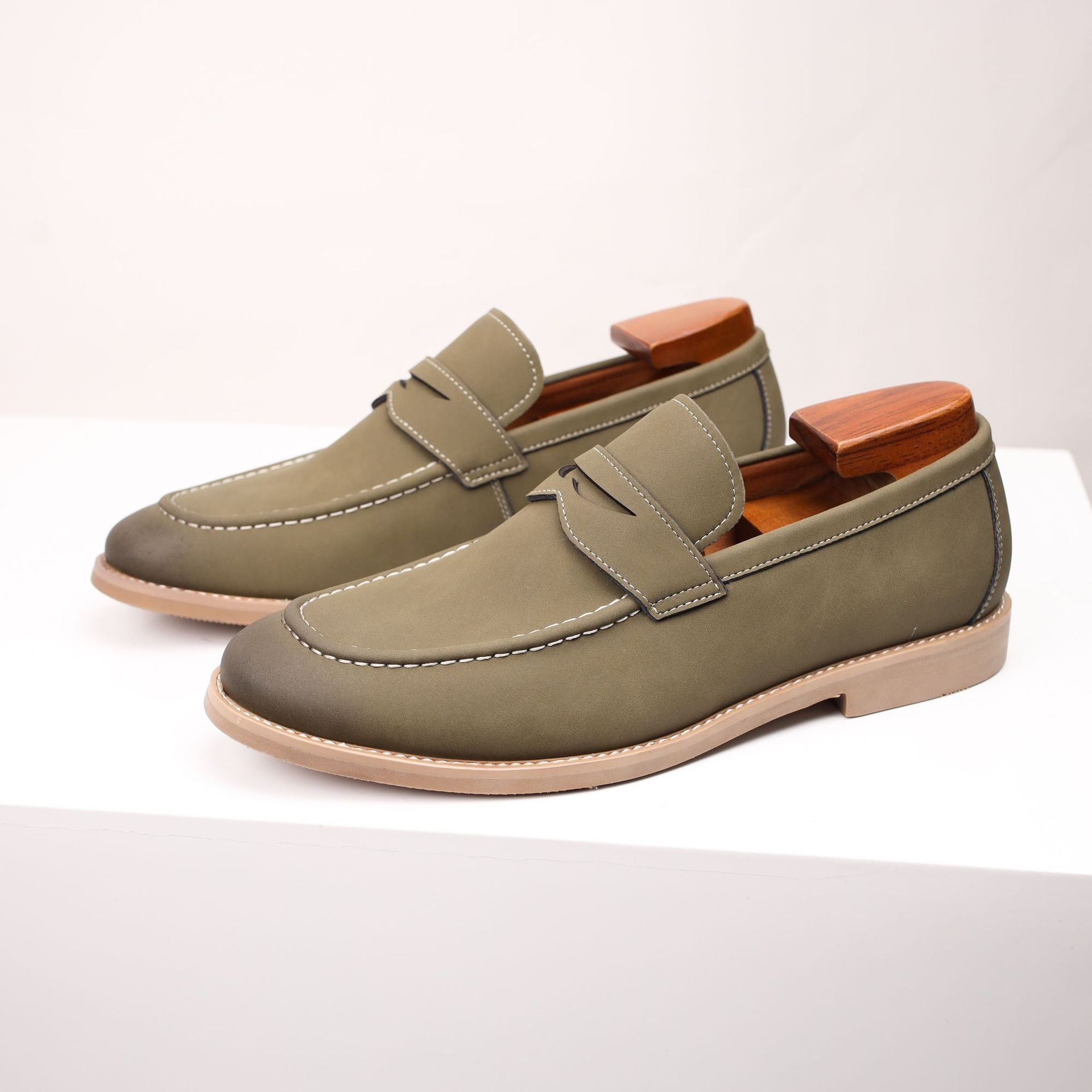 Pair of olive green leather penny loafers with wooden shoe trees, standing on a white pedestal.