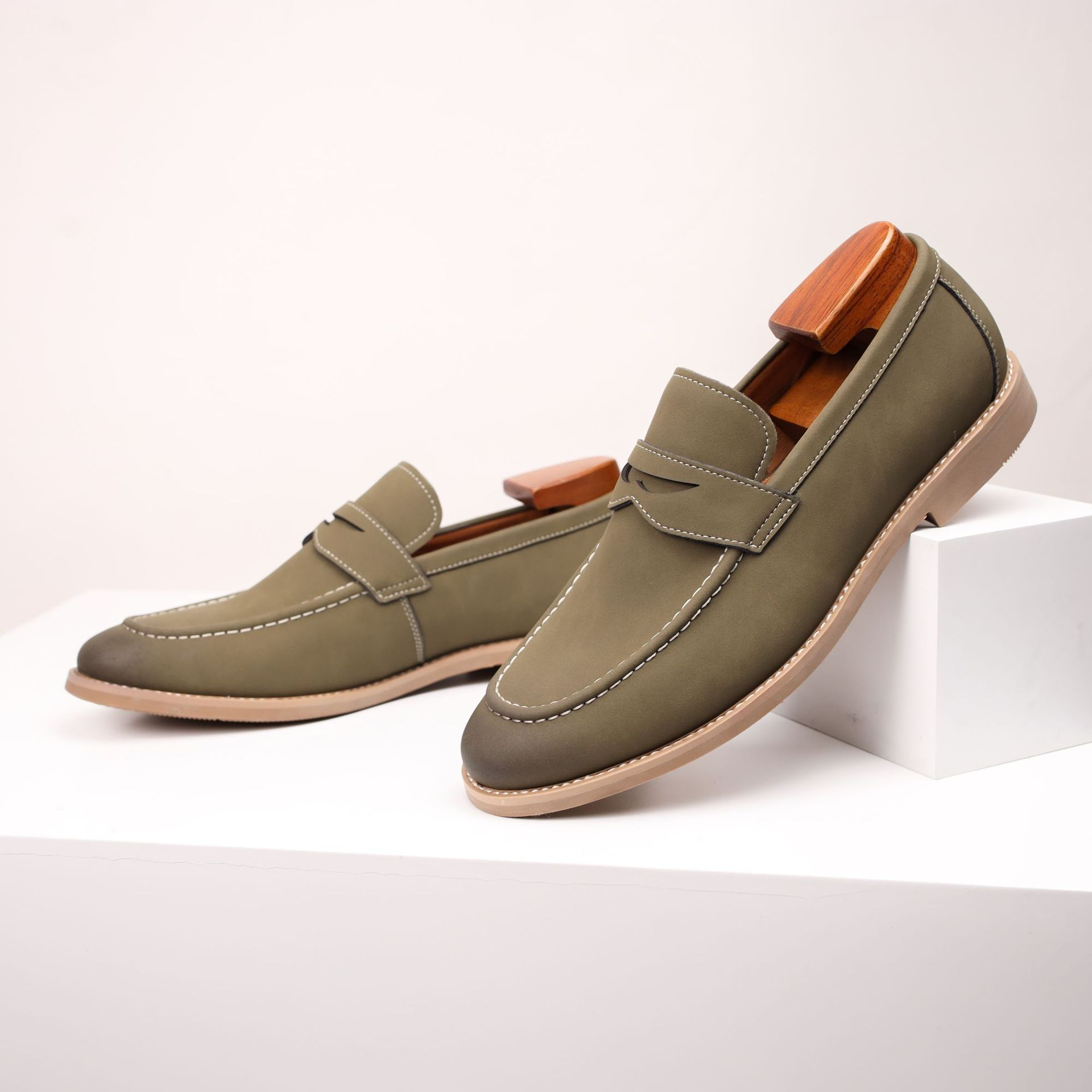 Pair of olive green suede penny loafers with wooden shoe trees, one leaning against a white platform block.