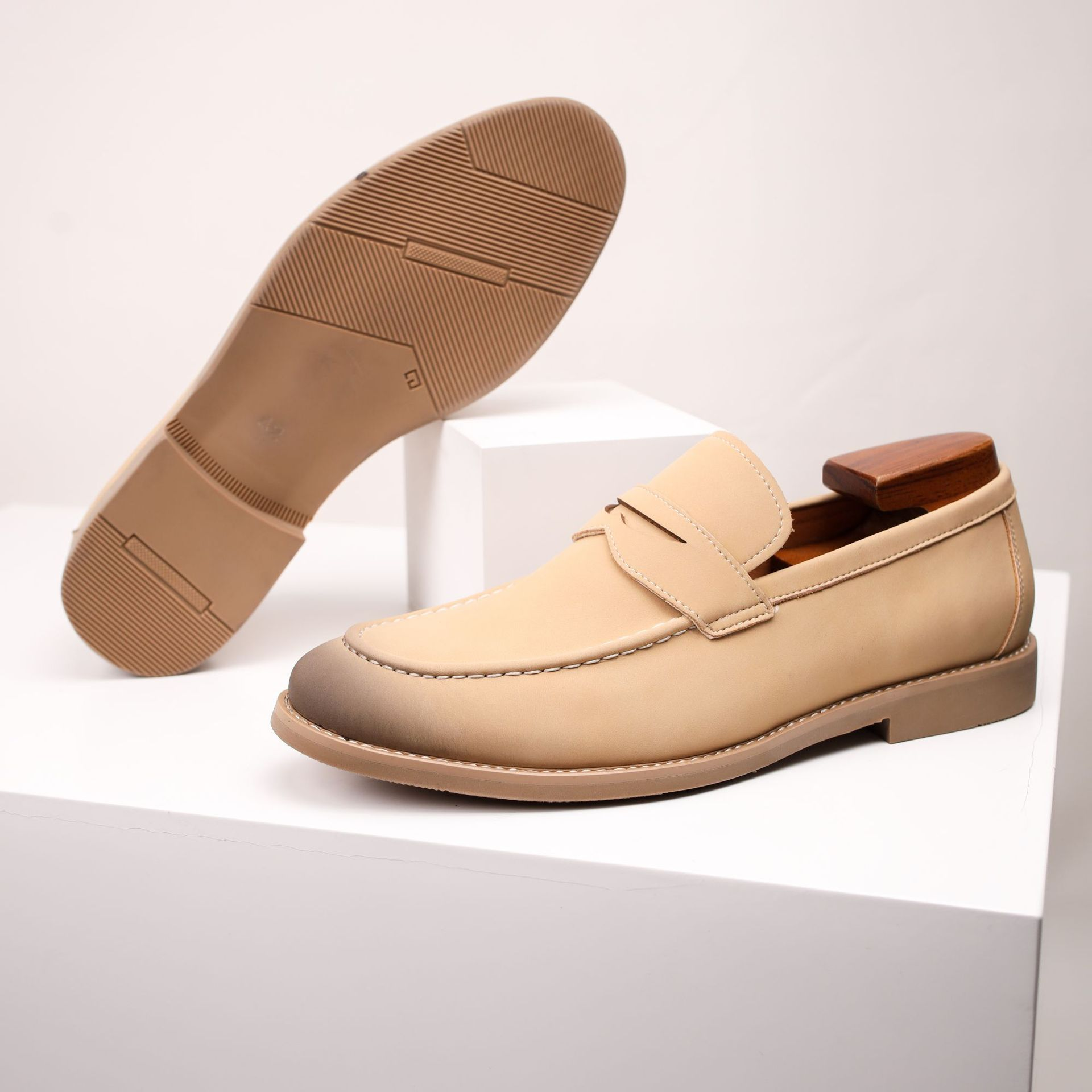 A tan leather penny loafer with a burnished toe, tilted on its side to show the tread sole, on a white pedestal.