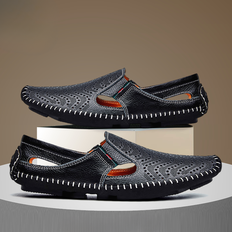 Pair of black perforated leather slip-on sandals with hand-stitched trim, one shoe stacked on a white display block above the other.
