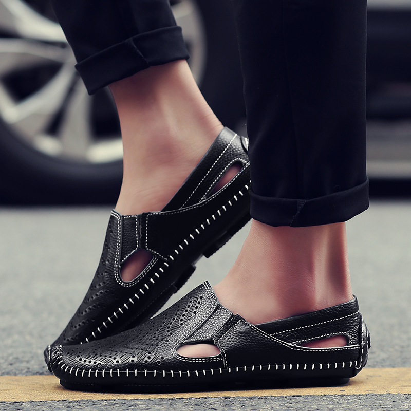Close-up of a model's feet wearing black perforated leather slip-on sandals with hand-stitched trim while standing outdoors on pavement.