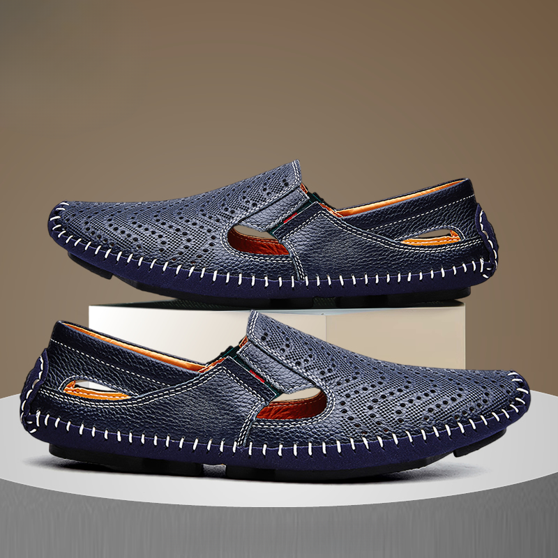 Pair of navy blue perforated leather slip-on sandals with hand-stitched trim, one shoe stacked on a white display block above the other.