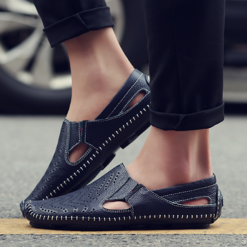 Close-up of a model's feet wearing navy blue perforated leather slip-on sandals with hand-stitched trim while standing outdoors on pavement.
