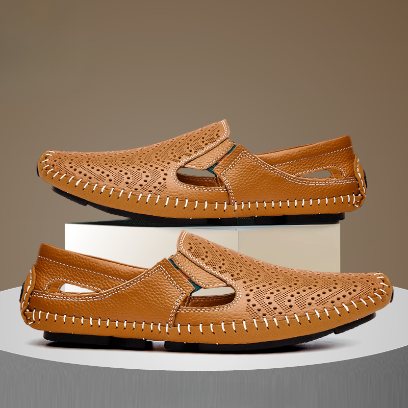 Pair of brown perforated leather slip-on sandals with hand-stitched trim, one shoe stacked on a white display block above the other.